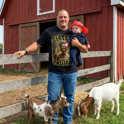 "Zombie Chef" black t-shirt with cannibal gourmet graphic and classic chillers logo; front view; on model with baby and goats in front of red barn.
